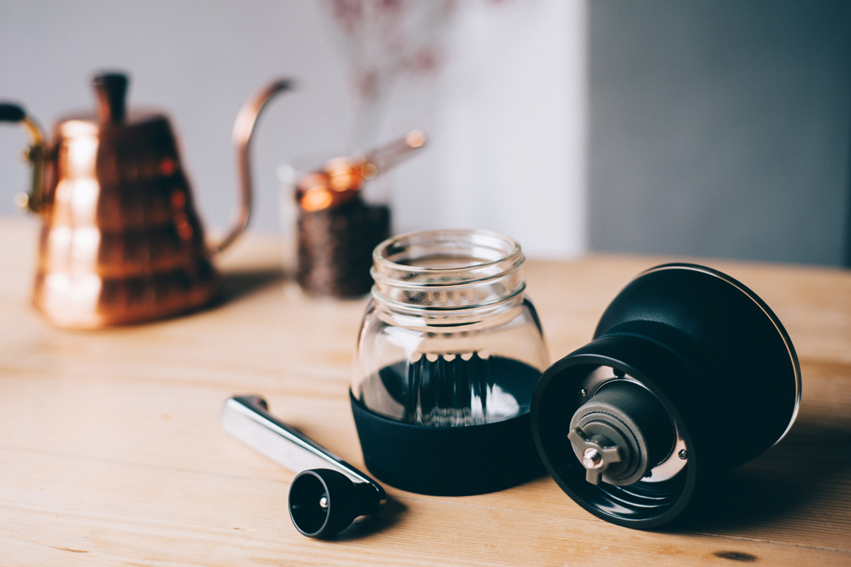 Moinho Skerton Pro da Hario desmontado com chaleira e solher de cobre no fundo. Em cima de uma mesa de madeira.